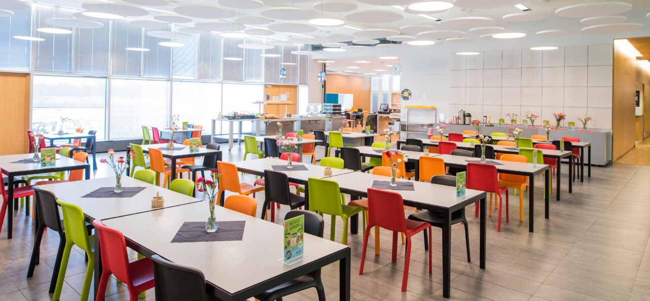 Ruokala, jossa monta pitkää ruokapöytää, jonka kummallakin pitkällä sivulla aina neljä tai kaksi värikästä tuolia. Pöydillä pieniä pöytäliinoja ja kukkamaljakoita sekä ravintolaesitteitä. Takana isot ikkunat, joiden edessä ruokalinjasto.